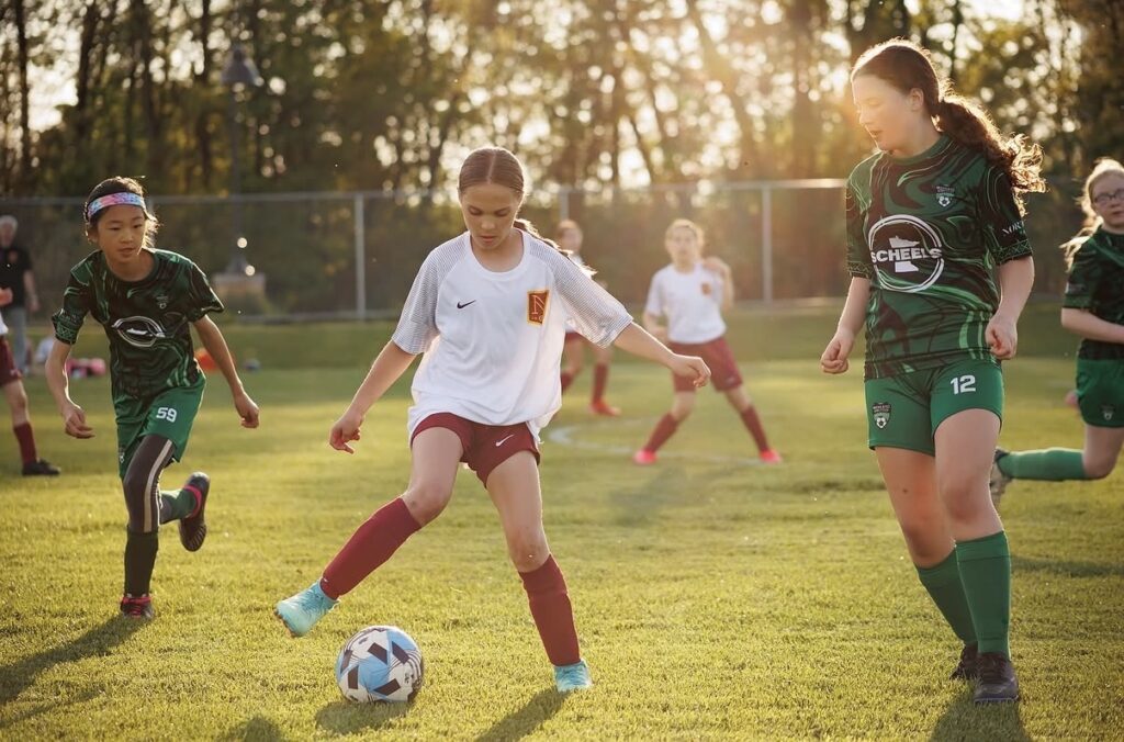 A girl kicks a soccer ball with other players surrounding her during a game